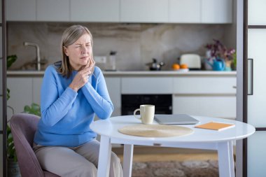 Diş ağrısından rahatsız olan olgun bir kadın mutfak masasında otururken elini yanağına dayayıp sağlık sorunlarını ve diş bakımı ihtiyacını sembolize ediyor.