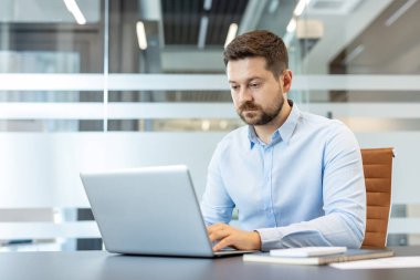 Çağdaş bir kurumsal çalışma alanındaki bir masada dizüstü bilgisayar kullanarak sakallı bir adam serbest veya iş görevlerinde aktif olarak çalışıyor.