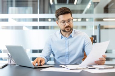 Gözlüklü ve mavi gömlekli iş adamı veri analizi yapıyor, önemli evrakları inceliyor, ve bilgisayarda daktilo yazıyor, verimlilik gösteriyor ve modern bir şirket işyerinde odaklanıyor.
