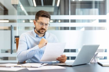 Gözlüklü ve açık mavi gömlekli bir iş adamı. Bir dizüstü bilgisayarla masada oturmak. Çağdaş bir ofis ortamında belge okumaya ve önemli şirket evraklarını incelemeye odaklanmış.