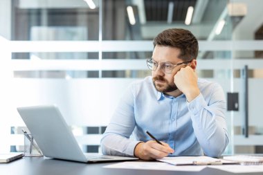 Gözlüklü bir işadamı ofis masasında oturuyor, yoğun bir çalışma sırasında kalemle yazarken kağıtlarla ilgili verileri dikkatlice değerlendiriyor, çözümler ve gelecekteki projeler için strateji geliştiriyor.
