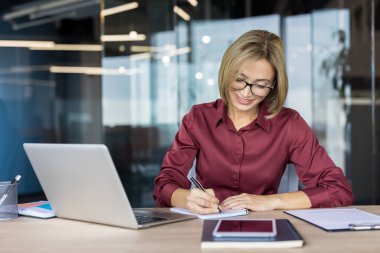 Gözlüklü, modern masasına gülümseyen kendine güvenen bir iş kadını, dizüstü bilgisayarlı ve akıllı telefonlu bir not defterine notlar yazıyor, organize ve işine odaklanmış.