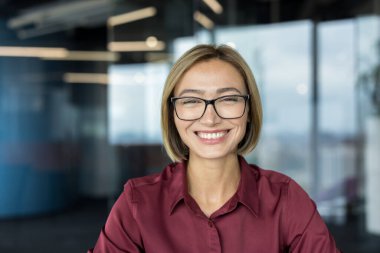 Kestane rengi gömlekli ve modern gözlüklü profesyonel iş kadını, çağdaş bir ofiste kameraya güvenle gülümseyen, liderlik, başarı ve yaklaşımı tasvir eden