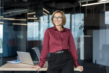 Çağdaş bir ofiste duran genç bir iş kadını, kendinden emin bir şekilde gülümseyen ve başka tarafa bakan, profesyonellik, liderlik ve kariyerinde başarı gösteren,