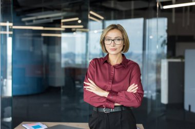 Kollarını kavuşturmuş profesyonel bir kadın yönetici, modern cam duvarlı bir ofiste kameraya güvenle bakıyor, liderlik, hırs ve kurumsal başarıyı iletiyor.