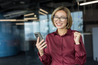 Profesyonel başarıyı kutlayan gülümseyen iş kadını kameraya bakar ve akıllı bir telefon tutar, modern ofis ortamında zafer ve neşe ifade eder.