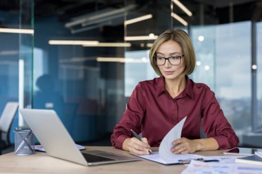 Masasında dizüstü bilgisayar kullanırken önemli belgelere odaklanan becerikli bir iş kadını, çağdaş bir şirket ortamında kendini adamışlık ve verimlilik gösteriyor.
