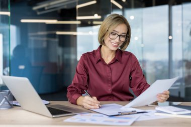 Parlak, modern bir çalışma alanındaki belgeleri inceleyen ve yazan mutlu bir iş adamı. Çalışmalarıyla aktif olarak ilgilenen ve veri gösteren bir dizüstü bilgisayar.