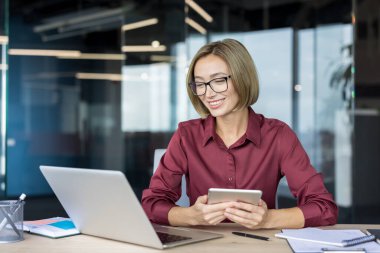 Modern bir ofiste çalışırken gülümseyen, dijital tablet ve dizüstü bilgisayarla birden fazla iş yapan, üretkenliği, yeniliği ve mesleki başarıyı simgeleyen kendine güvenen bir iş kadını.