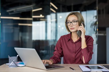 Akıllı bir telefonla konuşan kendine güvenen bir iş kadını aynı anda bir dizüstü bilgisayarda yazarak profesyonel ofis ortamında birden fazla iş gücü ve etkili iletişim göstermektedir.