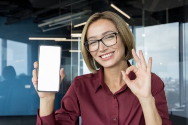 Gülümseyen iş kadını, gözlüklü, beyaz ekranlı bir cep telefonu sunarken, modern ofis ortamında başarılı ve onaylı bir jest yapıyor.