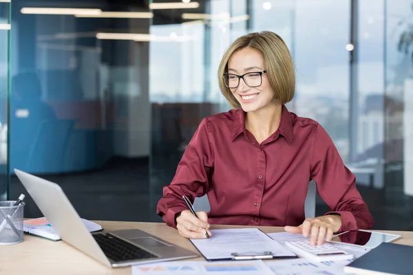 Modern ofisteki genç profesyonel kadın dizüstü bilgisayar, hesap makinesi ve not kullanırken gülümsüyor, masasında üretken ve kendinden emin finans ve muhasebe görevlerine odaklanıyor.
