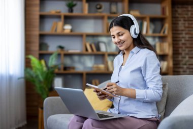Kulaklık takan, gülümseyen, e-öğrenen ya da uzaktan eğitim alan bir kadın ev ofisinden ya da oturma odasından dizüstü bilgisayar kullanarak not alıyor.