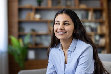 Uzun siyah saçlı, mavi tişörtlü genç bir Hintli kadın. Gülümsüyor ve gözlerini kaçırıyor. Arka planda kitap rafları olan modern bir ofis ortamını dikkatle dinliyor.