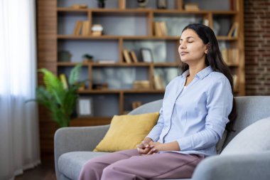 Kanepede gözleri kapalı oturan bir kadın, sakince nefesine odaklanıyor ve huzurlu bir ev ortamında zihinsel sağlığını ve duygusal dengesini korumaya çalışıyor.