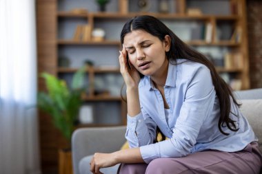 Kanepede oturan genç bir kadın, şiddetli baş ağrısı ya da migren ağrısı çekiyor, elini şakağına bastırılmış bir yüz ifadesiyle tutuyor, aile ortamında kendini iyi hissetmiyor.
