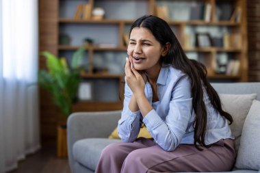 Kanepede yanağını acı içinde tutan genç bir kadın. Evde dişleri ağrıdığı için yüzünü buruşturuyor. Acil diş bakımı ve yardıma ihtiyacı var.