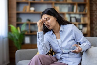 Şiddetli migren ve göz yorgunluğu çeken bir kadın, elinde gözlük tutan ve evinde aşırı çalışmaktan yorgun ve stresli bir şekilde kanepede otururken şakaklarını ağrıtan bir kadın.