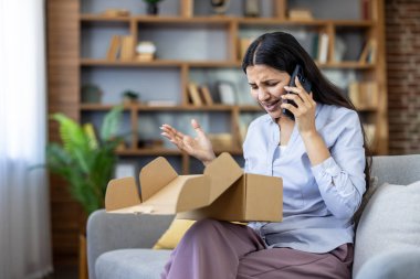 Kanepede oturan genç bir kadın, müşteri desteği için imdat çağrısı yapıyor, son teslimatıyla ilgili bariz bir sorunu olan açık bir karton kutuyu tutarken hayal kırıklığı ve hayal kırıklığı yaşıyor.