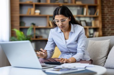 Gözlüklü genç Hintli kadın, mali durumları analiz ediyor, harcamaları yönetiyor ve modern ev ofisinde bir kanepede otururken dizüstü bilgisayar ve hesap makinesi kullanarak vergi raporları hazırlıyor.