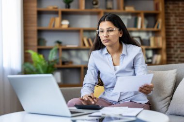 Kanepede oturan gözlüklü genç bir kadın, dizüstü bilgisayarla, belgelerle ve hesap makinesiyle harcamaları hesaplıyor, kişisel mali durumları ya da rahat ev ofisinden uzak işleri yönetiyor.