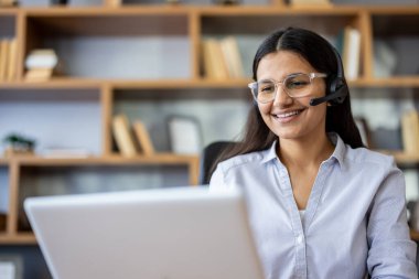 Profesyonel müşteri yardımı sağlayan kulaklık ve gözlük takan gülümseyen bir kadın, ev ofisinden uzaktan telefon ve internet iletişimi için dizüstü bilgisayar kullanarak çalışıyor.