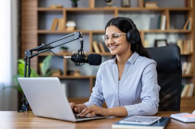 Kulaklık takan ve gözlük takan gülümseyen bir kadın stüdyo mikrofonu ve dizüstü bilgisayar kullanarak podcast kaydederek ev stüdyosundan veya ofisinden dijital ses içeriği yaratıyor.