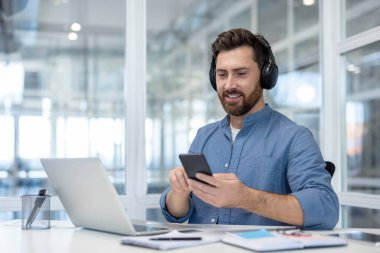 Kulaklık takan ve sakallı genç bir adam modern bir ofis masasında oturuyor, akıllı bir telefonla konuşurken gülümsüyor ve yanında modern bir çalışmayı temsil eden bir dizüstü bilgisayar var.