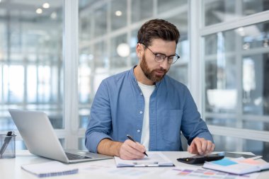 Modern ofis ortamında bir panoya yazı yazarken hesap makinesi ve dizüstü bilgisayar kullanarak, bütçe hesaplama ve raporlama üzerine yoğunlaşarak, gözlüklü bir adam.