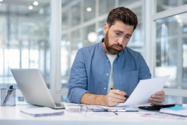Modern bir ofiste çok çalışan yorgun bir adam, masasında iş belgelerini düzenlerken stres ve patinaj yaşıyor, bunaltıcı görevlere ara vermesi gerekiyor.