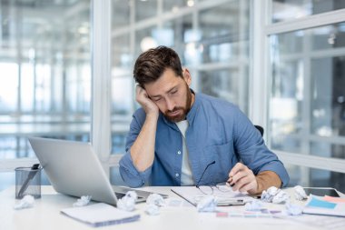 Ofis masasında elinde gözlük tutan yorgun bir adam. Etrafı buruşmuş kağıtlar ve dizüstü bilgisayarlarla çevrili. Baskı altında bitkinlik, yorgunluk ve hüsranı gösteriyor.
