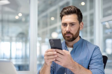 Ciddi, yakışıklı, profesyonel, cep telefonu tutan, mesaj okuyan ya da internette gezinen, modern bir ofiste çalışırken müşteri ya da meslektaşlarıyla iletişim kuran bir erkek.