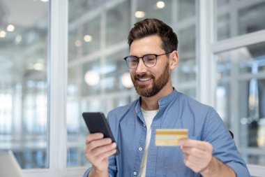 Adam elinde bir kredi kartı ve akıllı telefon tutarken gülümsüyor, uygun dijital ödeme için evden veya ofisten online alışveriş veya mobil bankacılık işlemleri yapıyor