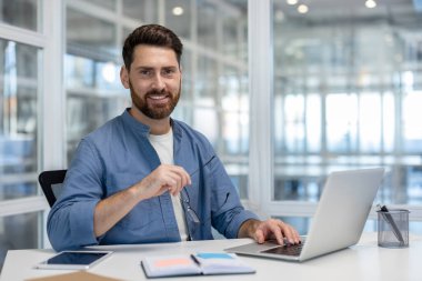 Kendine güvenen sakallı bir adam, parlak bir masada dizüstü bilgisayarla çalışırken kameraya gülümsüyor, profesyonelliği, girişimciliği ve modern bir çalışma alanında üretkenliği somutlaştırıyor.