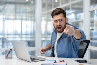 Modern bir ofisteki bir masadan seyirciye bağıran ve doğrudan işaret eden bir adam, şirket işyeri ortamında öfke, hayal kırıklığı ve yüzleşme gösteriyor.