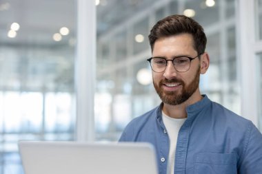 Dizüstü bilgisayarla çalışırken gülümseyen, kendine güvenen sakallı bir adam. Çalışmayı, odaklanmayı, teknolojiyi ve profesyonel ofis ortamında başarıyı temsil ediyor.