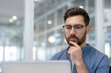 Gözlüklü ve rahat gömlekli düşünceli bir adam modern bir ofiste dizüstü bilgisayara odaklanıyor, verileri analiz ediyor ve sessizce çözümleri planlıyor.