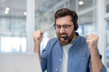 Kulaklık takan ve gözlüklü bir adam kalabalık bir ofiste dizüstü bilgisayarına yumruk atıyor ve bağırıyor. İş baskısı, teslim tarihi ve teknik arıza yüzünden stres ve hüsranla dolup taşıyor.