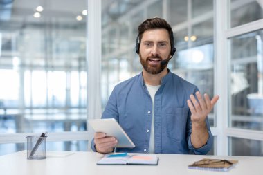 Kulaklık takan sakallı bir adam, elinde dijital bir tablet, ve bir video çağrısına katılırken el kol hareketi yapıyor, uzaktan çalışmayı ve profesyonel iletişimi temsil ediyor.