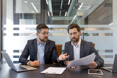 Takım elbiseli iki işadamı modern cam duvarlı bir toplantı odasında doküman ve dizüstü bilgisayar üzerinde işbirliği yapıyor, stratejiyi tartışıyor, verileri analiz ediyor ve sonraki adımları birlikte planlıyorlar.