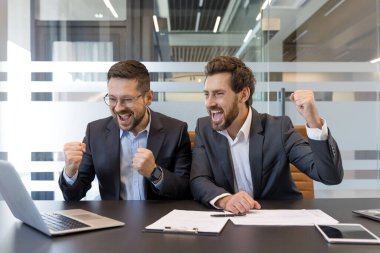 İki erkek iş ortağı tezahürat yapıyor ve yumruklarını kaldırıyor. Dizüstü bilgisayara bakıyor. Bir zaferi veya başarıyı ofis masasındaki bir toplantıda kutluyor.