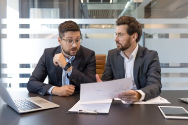 Takım elbiseli iki erkek işadamı modern bir ofis masasında mali rapor kağıtlarını tartışıyor, bir toplantı sırasında iş stratejisi planlarken pazar verilerini işbirliği yapıyor ve analiz ediyorlar.