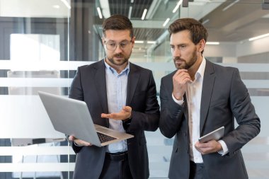 Bir dizüstü bilgisayarda veri analizi yapan ve işbirliği yapan iki profesyonel işadamı, odaklanmış bir tartışmanın ortasında çağdaş bir ofis ortamında duruyorlar.