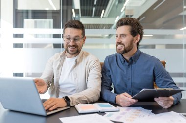 İki erkek iş adamı modern bir ofiste ortak bir toplantı yapıyorlar, biri dizüstü bilgisayarı işaret ederken diğeri ise hem gülümseyen hem de tartışan bir dosya tutuyor.