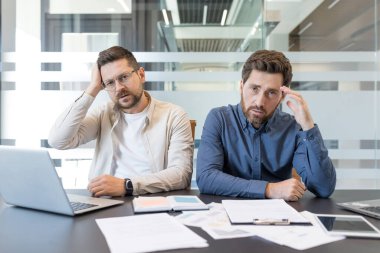 İki erkek girişimci bir ofis masasında oturmuş, stres dolu ifadeler ve başlarının üstünde ellerle kameraya bakıyor, tükenmişlik ve iş başarısızlığı altında eziliyorlar.