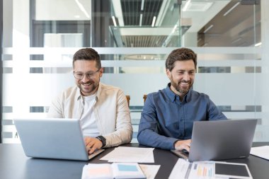 Parlak, modern bir ofis masasında iş birliği yapan, dizüstü bilgisayarlar ve belgeler üzerinde çalışan, odaklanmış, takım çalışması ve pozitif enerji içeren bir iş projesi planlayan iki gülümseyen erkek arkadaş.