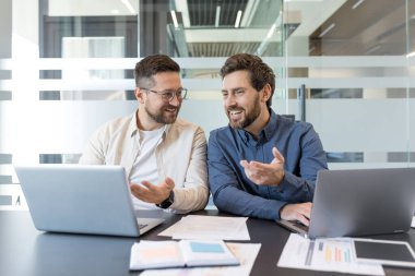 İki gülümseyen erkek iş arkadaşı, çağdaş ofis ortamında dizüstü bilgisayarlar ve belgelerle çalışırken aktif olarak fikirleri tartışıyor ve içgörülerini paylaşıyorlar.