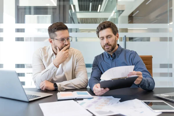 Deneyimli iş adamları kurumsal mali raporları analiz ediyor, önemli belgeleri inceliyor ve modern bir ofiste resmi bir iş toplantısında yeni bir proje sözleşmesini tartışıyorlar.