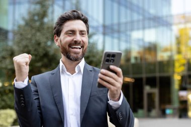 Resmi giyinmiş genç, mutlu, sakallı iş adamı dışarıda takılıyor. Akıllı telefonuna büyük bir gülümsemeyle bakarken zafer jestini kutluyordu. Çevrimiçi başarı ve olumlu haberleri temsil etmek