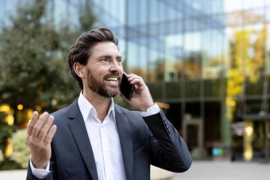 Takım elbiseli gülümseyen işadamı, modern cam ofis binasının önünde telefonla konuşuyor, güvenli bir şekilde iletişim kuruyor ve anlaşmaları tartışırken aynı zamanda kentsel, profesyonel başarı ve fırsatların tadını çıkarıyor.